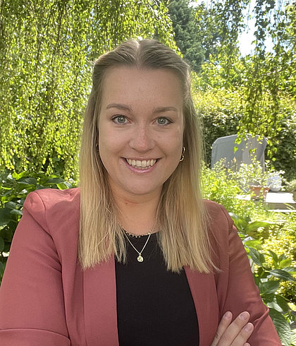 Lächelnde Frau mit blondem Haar, rosa Blazer und verschränkten Armen vor Garten